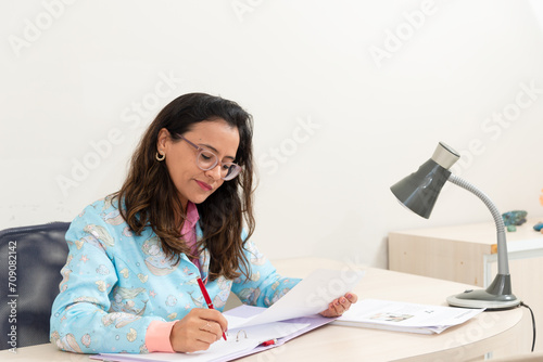 Portrait of a psychopedagogue in her office, wearing a colorful coat, sitting, making notes on paper. Helps with school performance. © Thales