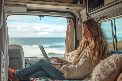 A beautiful woman in stylish clothes works with a laptop in a trailer, mobile home on the ocean. Freelancer. Work remotely. trailer home. Camping vacation