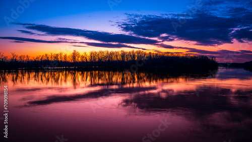Sunset with bare tree forest reflected on water and blazing orange-colored nightfall
