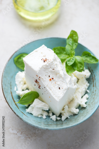 Turquoise bowl with a block of feta cheese and green basil, vertical shot on a light-beige stone background, selective focus