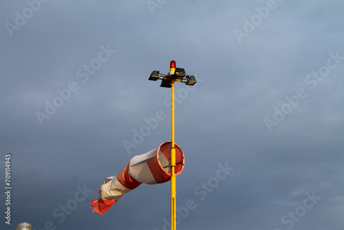 Windsock torn by the wind. Mast with red signal lights of the aerodrome against a dramatic stormy sky. Aerodrome aviation equipment at sunset against a dramatic stormy sky.