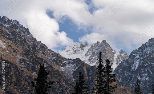 Snow-covered mountain ranges, cloud-draped peaks, fir trees at the base. Central Asia. Mountainous landscape. Autumn.