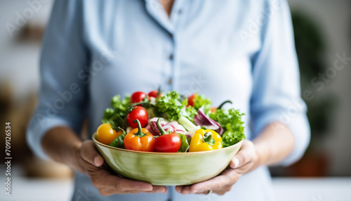 Aged woman smiling happily and holding a healthy vegetable salad bowl on blurred kitchen background  with copy space