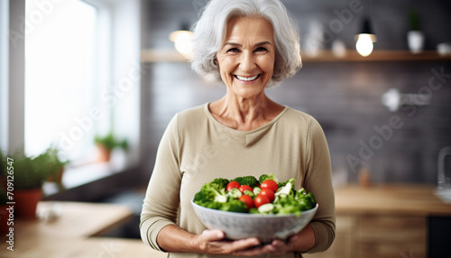 Aged woman smiling happily and holding a healthy vegetable salad bowl on blurred kitchen background, with copy space