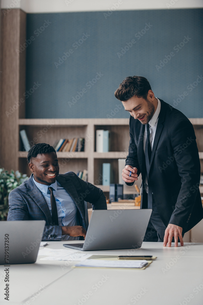 Two happy businessmen in formalwear using computer while working in the office together Two happy businessmen in formalwear using computer while working in the office together