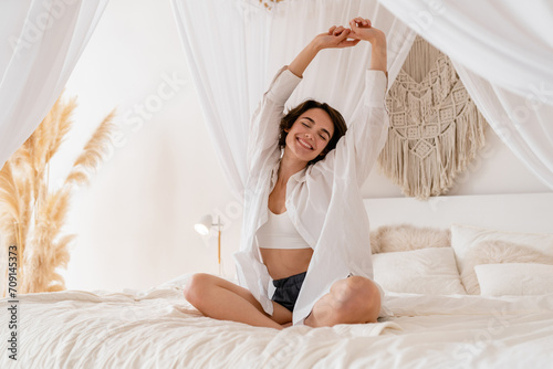 woman wake up in morning in boho style bedroom, sitting on bed lazy stretching, happy smiling
