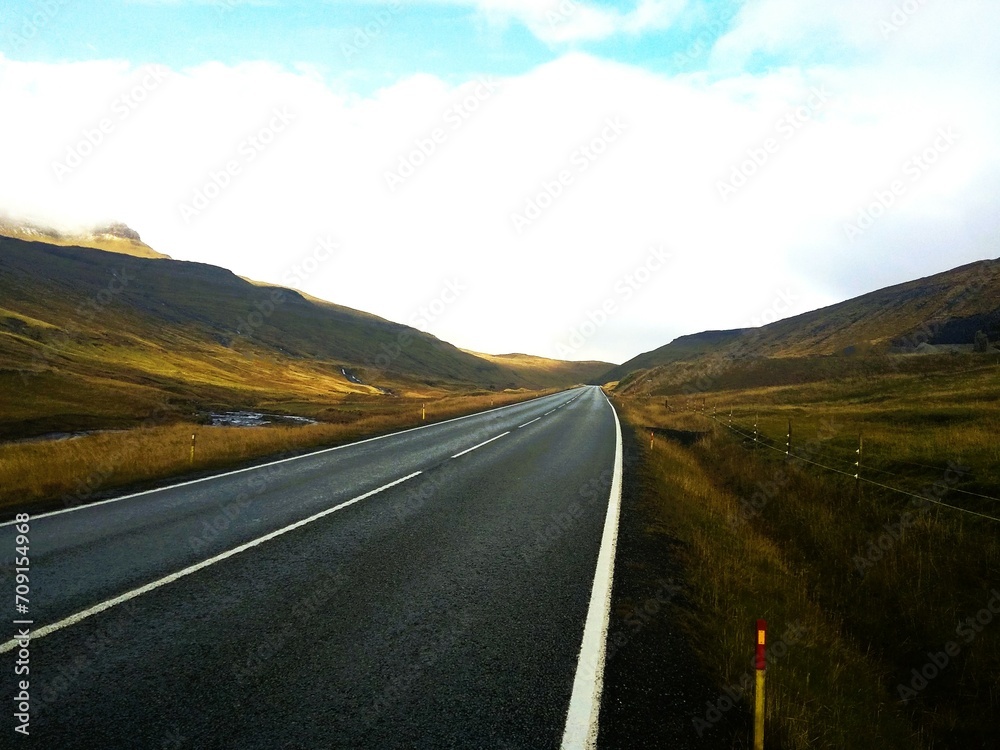 Droga asfaltowa rozciąga się pomiędzy górami. Górska droga znajdująca się na Wyspach Owczych. Surowy klimat widoczny w oddali. - obrazy, fototapety, plakaty 