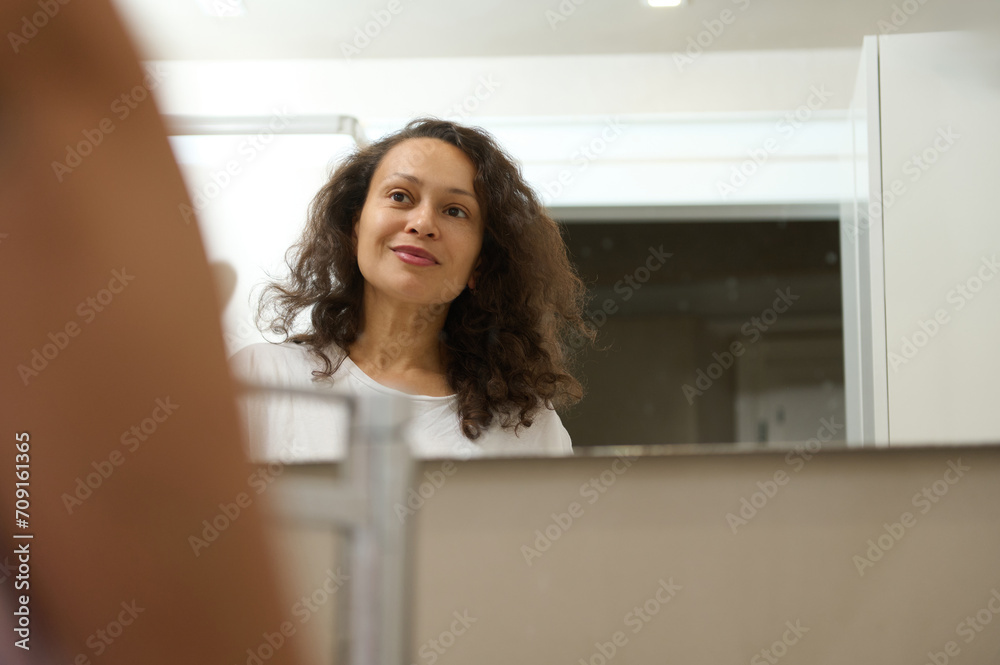 Reflection in the bathroom mirror of a multi ethnic smiling happy woman admiring her appearance. Home spa and beauty