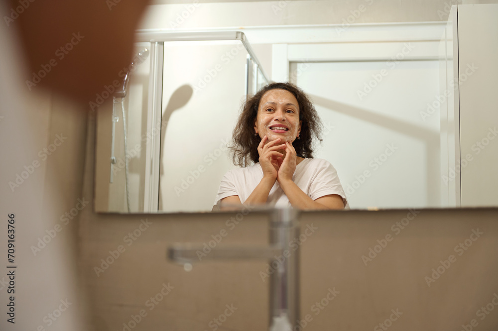 Reflection in mirror of pretty woman with radiant healthy skin, smiling looking at herself while doing spa procedures Reflection in mirror of pretty woman with radiant healthy skin, smiling looking at herself while doing spa procedures