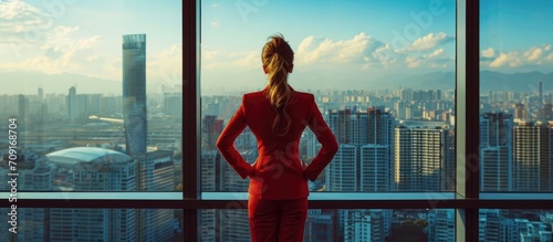 Confident female CEO managing company's investment strategy, wearing red suit in office, overseeing city view.