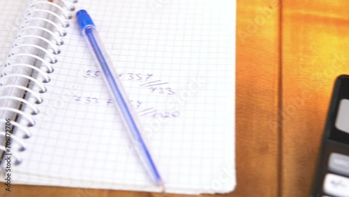 A calculator and a notebook on a brown wooden table under table lamp with warm light, slow motion video.
