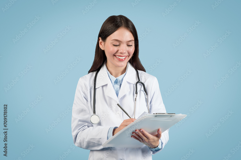 Happy woman doctor writing on clipboard against blue backdrop Happy woman doctor writing on clipboard against blue backdrop