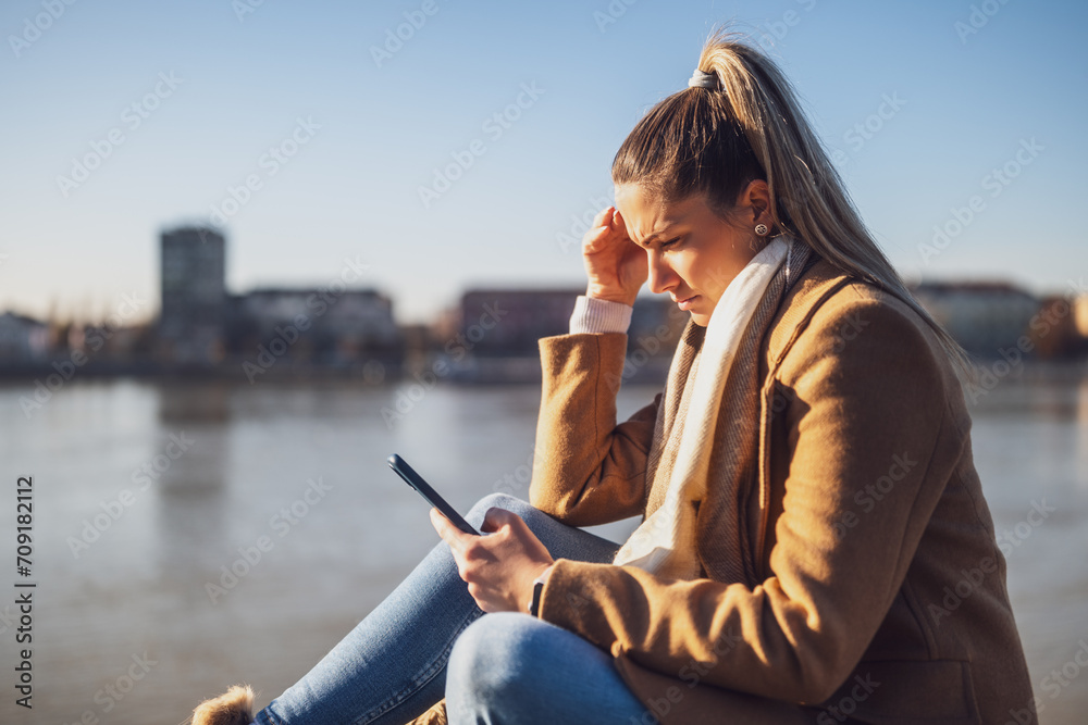 Sad woman holding phone while sitting by the river. Toned image.