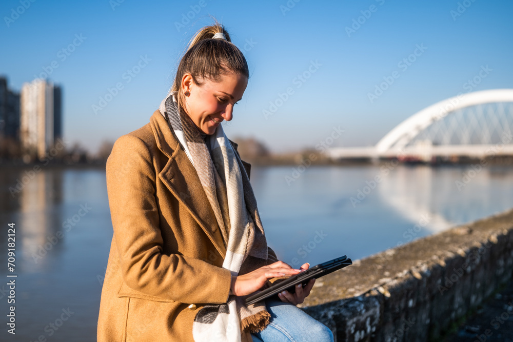 Beautiful woman in warm clothing using digital tablet and enjoys resting by the river on a sunny winter day.