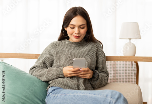 Cheerful young woman resting on couch, using smartphone