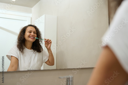 Reflection in the mirror of a happy confident good looking woman brushing teeth and smiling, admiring her appearance