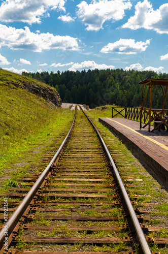 Railway, going far beyond the horizon; It passes through the flat terrain, along sides of fields and meadows with yellow and green hues, trees are also visible; The sky is cloudless color photo