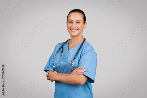 Happy nurse lady with folded arms posing wearing blue workwear and standing on gray studio background, smiling to camera photo