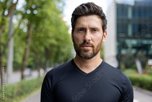 Confident bearded man in casual attire standing outdoors with a focused expression and urban backdrop