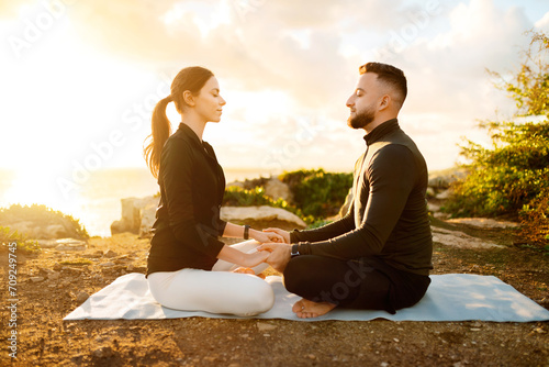 Focused couple holding hands in meditation on seaside cliff at dawn