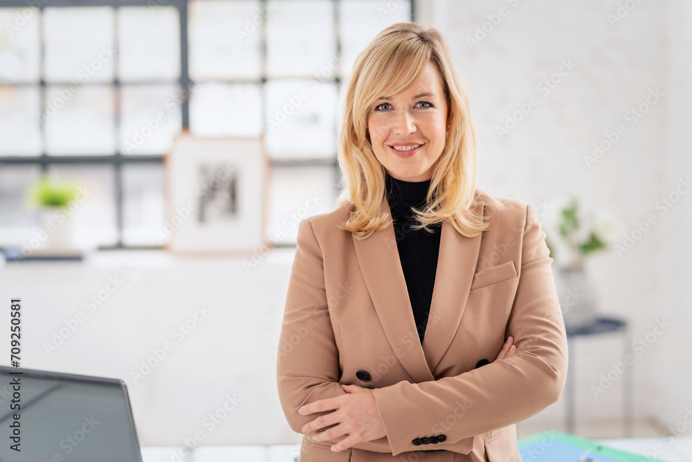 A middle-aged woman with blonde hair standing in the office. She is wearing a jacket and smiling into the camera.