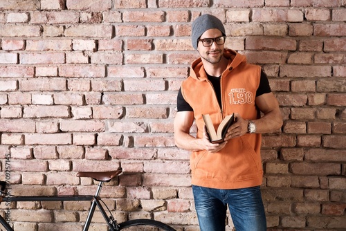 Young smart trendy caucasian man in glasses and cap standing at brick wall reading book. Copyspace for text. .