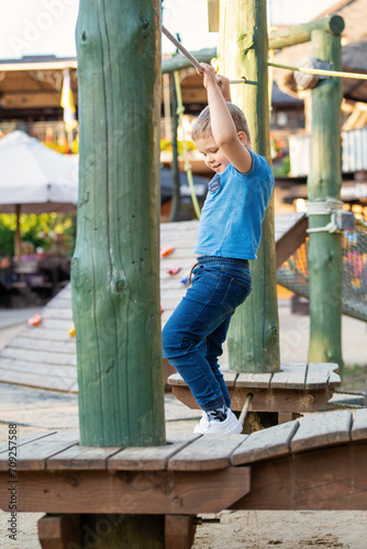 Smiling cute boy in blue clothes walking on obstacle course holding on to rope. © Linas T