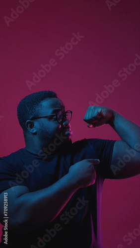 Showing power. Color light people. Strong confident man in glasses pointing at his biceps isolated on dark pink empty space background. photo