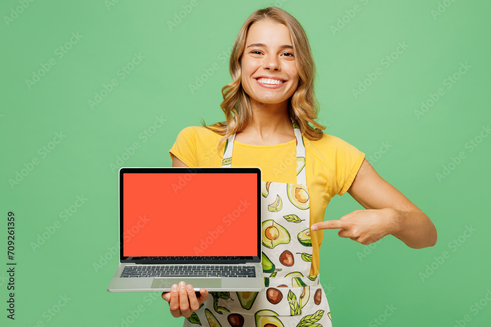 Young housewife housekeeper chef cook baker IT woman wear apron yellow t-shirt hold use work point on blank screen laptop pc computer recipe isolated on plain green background. Cooking food concept. Young housewife housekeeper chef cook baker IT woman wear apron yellow t-shirt hold use work point on blank screen laptop pc computer recipe isolated on plain green background. Cooking food concept.