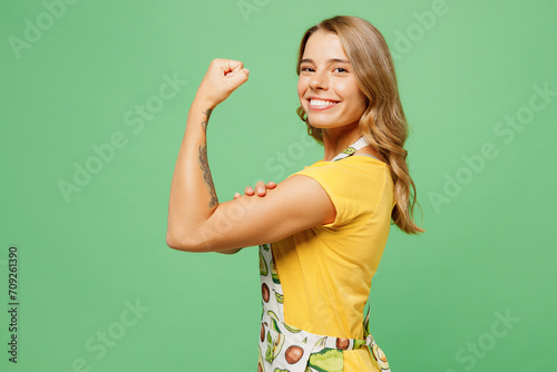 Side view young strong fun happy housewife housekeeper chef cook baker woman wear apron yellow t-shirt show muscles on hand look camera isolated on plain pastel green background. Cooking food concept. photo