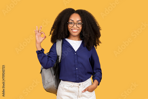 Happy black female student with backpack in glasses shows ok sign with hand isolated on yellow studio background photo