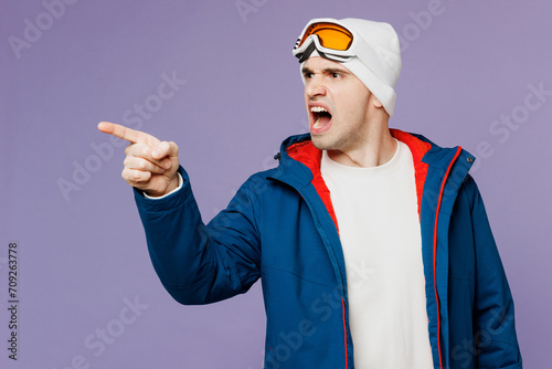Skier angry man wear warm blue windbreaker jacket ski goggles mask point finger aside scream shout spend extreme weekend winter season in mountains isolated on plain purple background. Hobby concept.