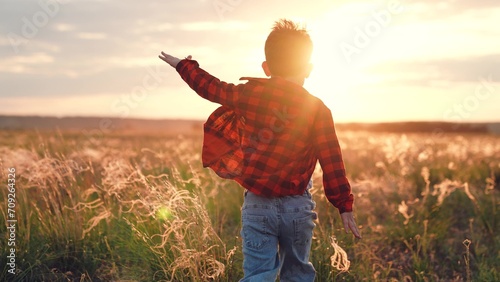 Positive boy runs along field with growing plants at sunset in autumn evening. Boy runs imitating flight with arms out to sides in rural nature with agriculture. Boy feels freedom running