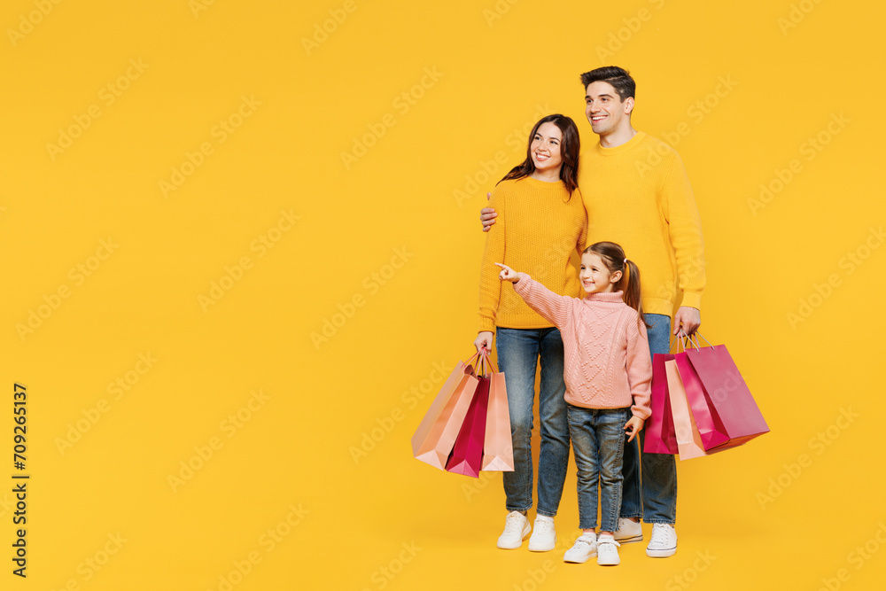 Full body young parent mom dad with child kid girl 7-8 years old wear pink sweater casual clothes hold shopping bags point aside isolated on plain yellow background. Black Friday sale buy day concept. Full body young parent mom dad with child kid girl 7-8 years old wear pink sweater casual clothes hold shopping bags point aside isolated on plain yellow background. Black Friday sale buy day concept.