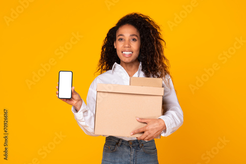 Black Woman Showing Smartphone Screen Holding Carton Box, Yellow Background