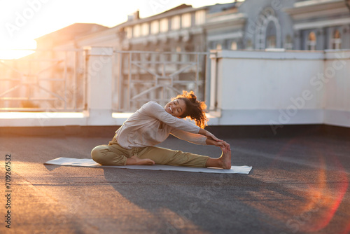 Young sporty happy mixed-race woman doing stretching exercises on yoga mat on house roof in morning