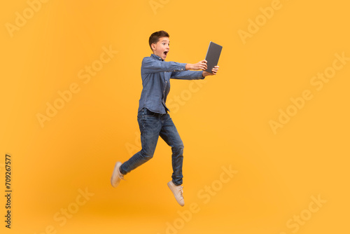 Surprised teenage boy jumping with digital tablet in hands over yellow background photo