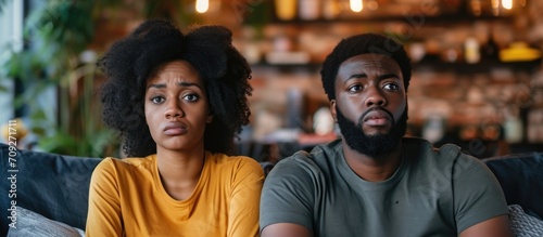 Concerned young African American couple watching TV, displaying skepticism and anxiety, upset due to a problem.