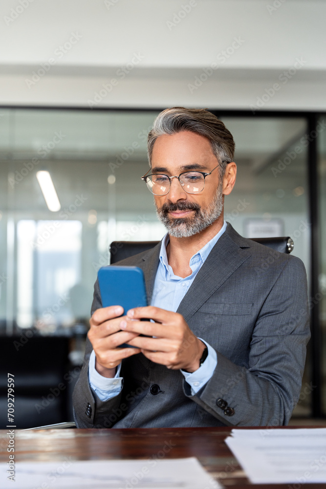 Vertical portrait of middle aged Hispanic business manager using cell phone mobile app. Serious Latin or Indian mature man businessman holding smartphone sit in office working online on gadget.