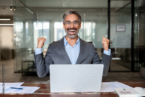 Middle-aged Latin or indian businessman screaming proud cheering with excitement , man read great news on smartphone celebrating victory and success sitting front of laptop computer working at office.