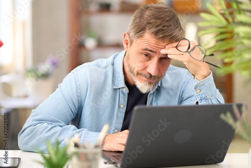 Casual mid adult man with laptop computer at desk in home office, having problem, troubled. Portrait of older gray haired bearded guy thinking. Businessman managing business on internet.   photo