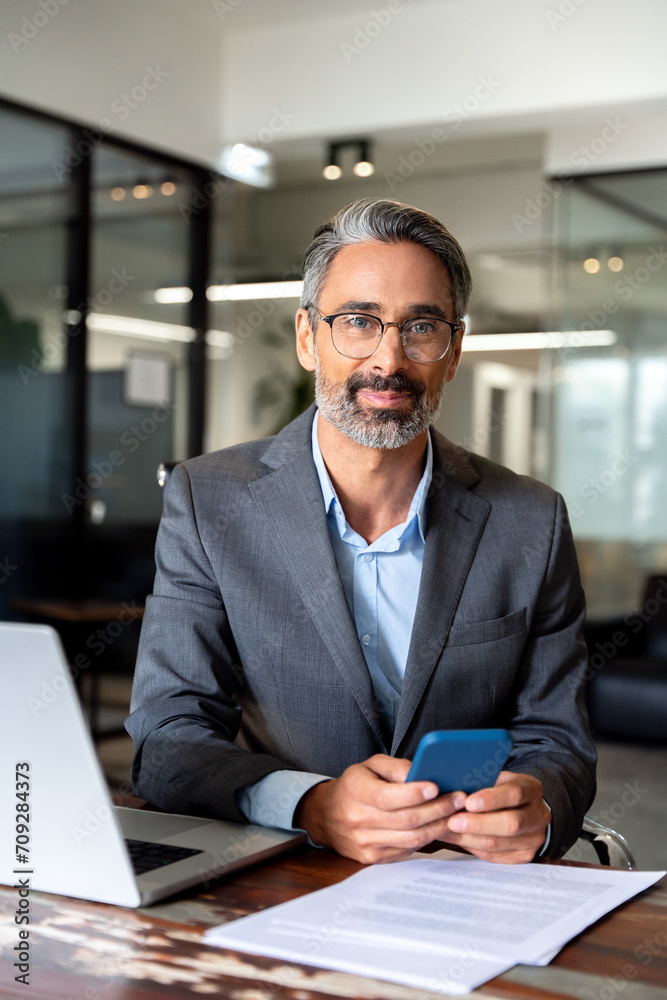 Vertical portrait of middle aged Hispanic business manager using cell phone mobile app. Smiling Latin or Indian mature man businessman holding smartphone sit in office working online on gadget. Vertical portrait of middle aged Hispanic business manager using cell phone mobile app. Smiling Latin or Indian mature man businessman holding smartphone sit in office working online on gadget.
