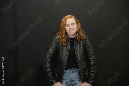 Young woman with red hair in black jacket and jeans poses in studio before a black background