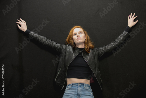 Young woman with red hair in black jacket and jeans poses in studio before a black background