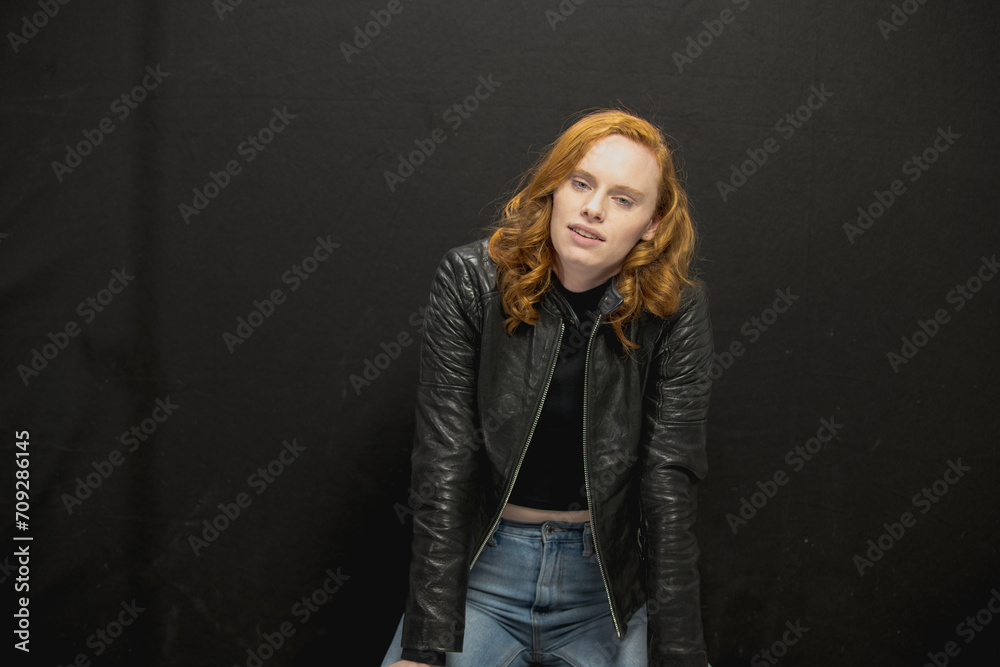 Young woman with red hair in black jacket and jeans poses in studio before a black background Young woman with red hair in black jacket and jeans poses in studio before a black background