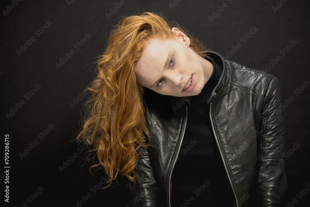 Young woman with red hair in black jacket and jeans poses in studio before a black background Young woman with red hair in black jacket and jeans poses in studio before a black background