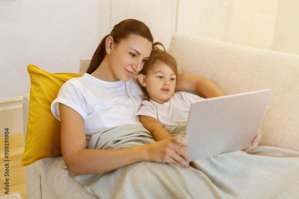 Mother and little daughter with digital tablet relaxing under blanket on sofa Mother and little daughter with digital tablet relaxing under blanket on sofa