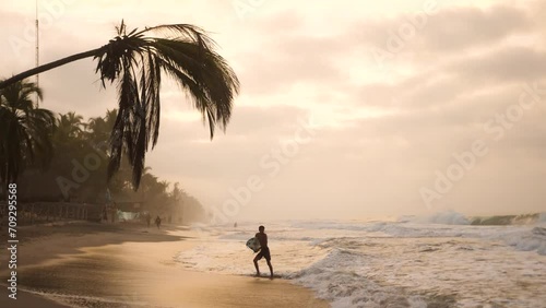 Surfer walking out of the sea on the Palomino beach at beautiful sunset, Colombia. Bent palm tree over the beach. Surfer part of Colombia, Palomino. photo