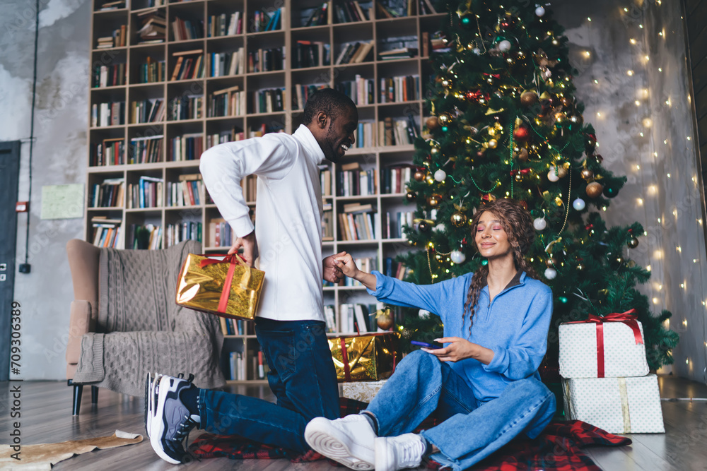 Cheerful African American man handing a golden gift to a seated Caucasian woman with a smartphone, by a Christmas tree in a book-laden interior, evoking a festive and loving holiday atmosphere