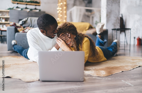 Wallpaper Mural African-American couple enjoying fun moment together while lying on floor in cozy living room, with laptop. woman, laughing partially covering her eyes, man is whispering something humorous to her Torontodigital.ca